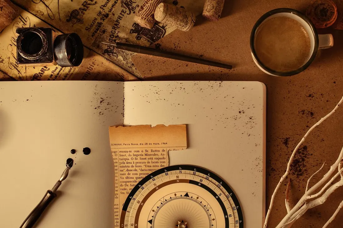 A vintage desk scene with an open journal, a compass dial, an ink pen, and a cup of coffee.