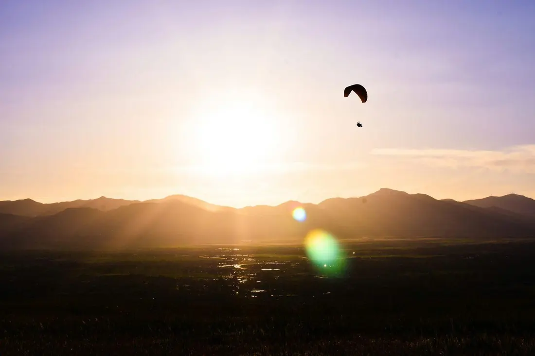 A paraglider is silhouetted against a glowing sunset sky above a mountain valley.