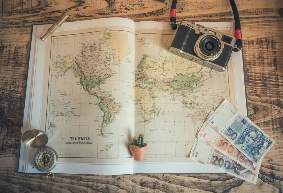 An open world map on a wooden table with a vintage camera, a compass, a small cactus, and foreign banknotes.
