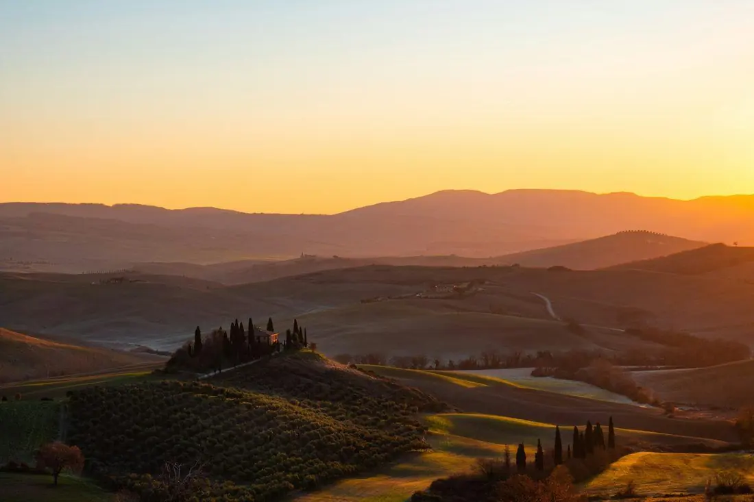 A hillside farmhouse ringed by cypress trees at sunrise, with rolling countryside fading into a warm orange sky.