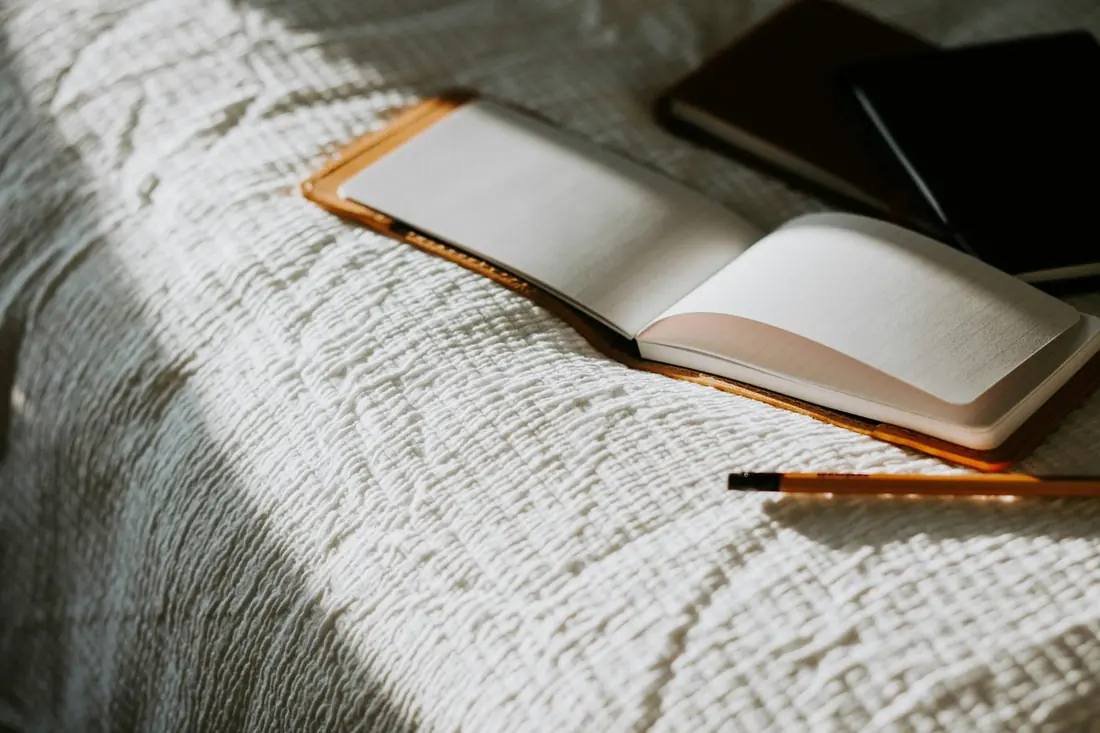 An open notebook and a pencil rest on a white linen bedspread in soft window light.