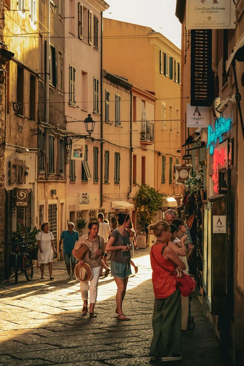 Travelers walk down a sunlit cobblestone lane lined with pastel buildings in late-afternoon light.