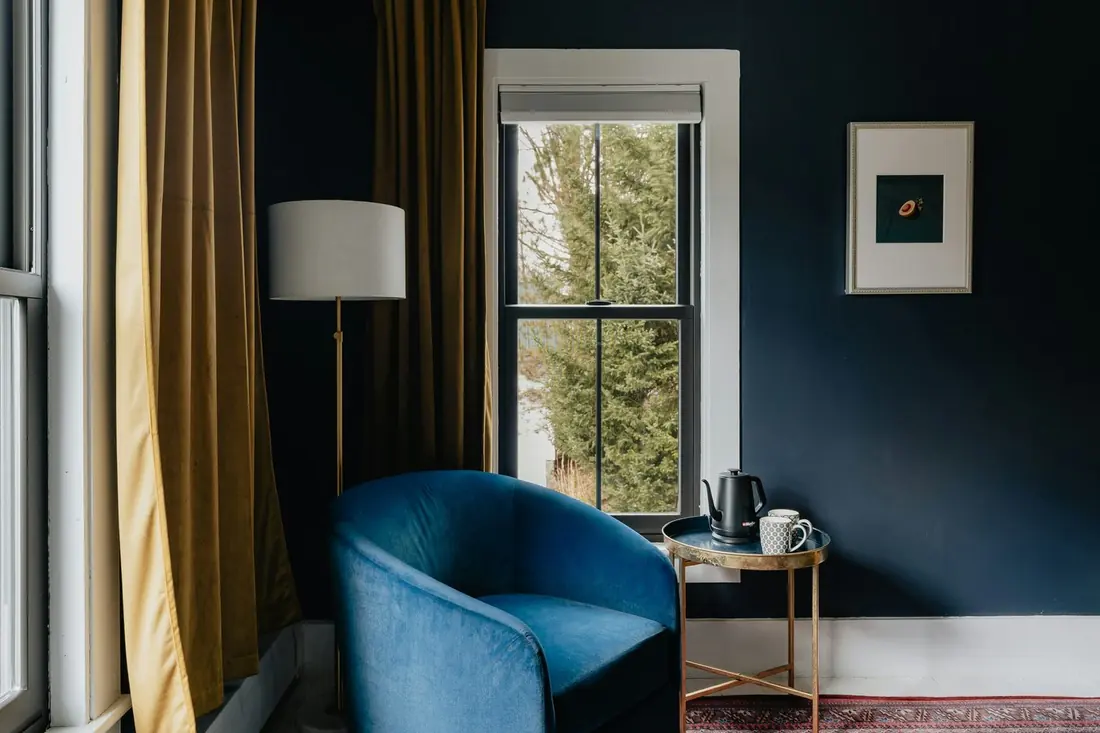 A blue velvet armchair in a navy-walled room beside a tall window, with a small brass side table.