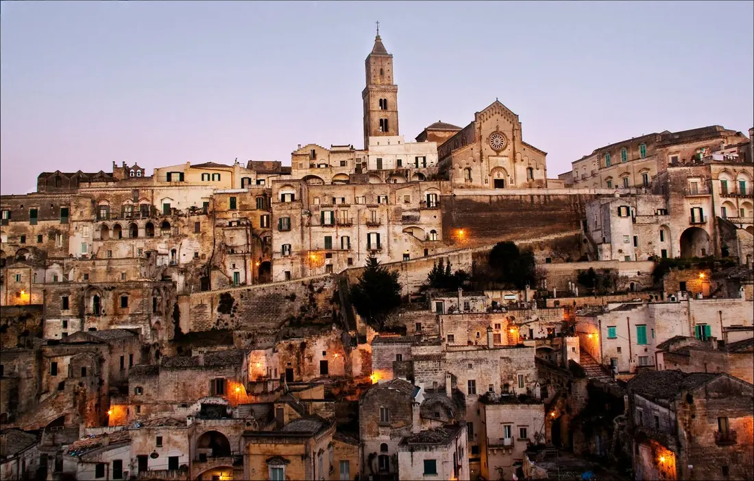 Matera's stone cathedral and hillside houses at blue hour, with warm lights coming on.