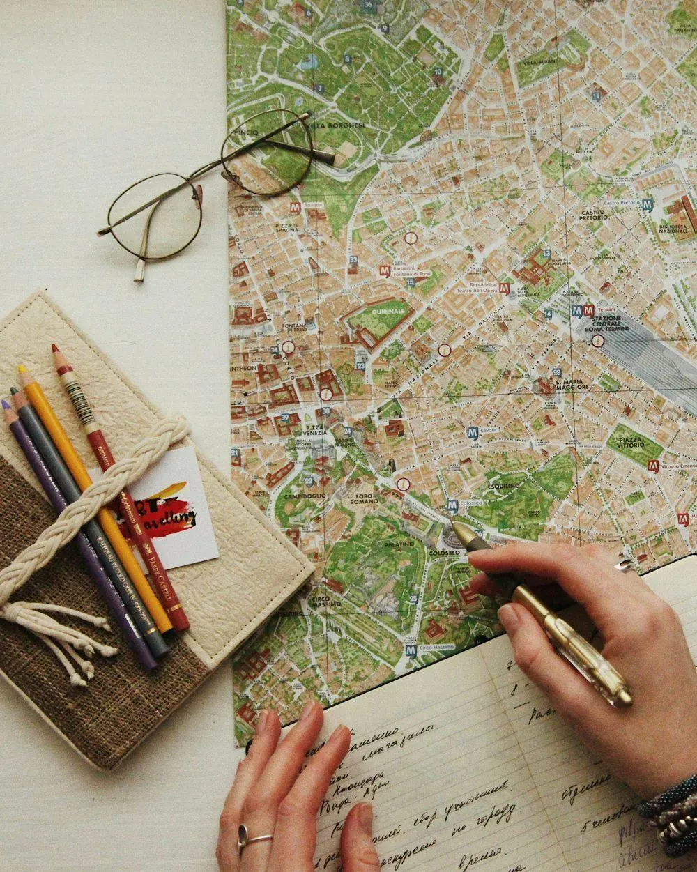 Hands mark a city map with a pen beside an open journal, reading glasses, and colored pencils.