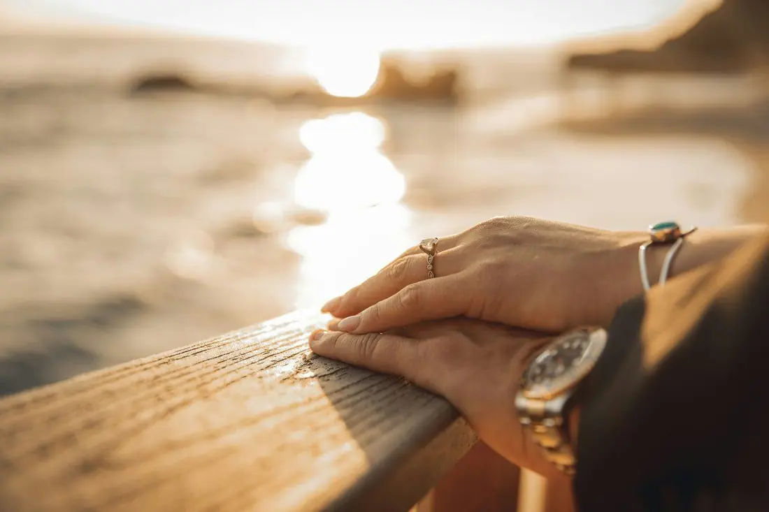 Hands rest on a weathered wooden ship rail overlooking water glittering in the low sun.