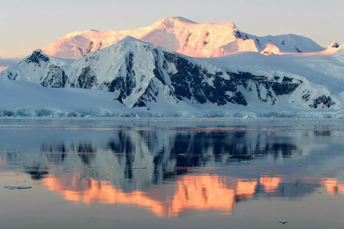 Snow-covered peaks glow pink at sunrise, reflected in the still polar water below.