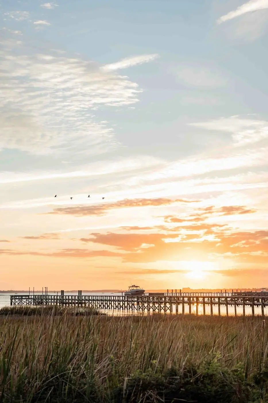 A long wooden pier stretches across calm water at sunrise, with marsh grass in the foreground.