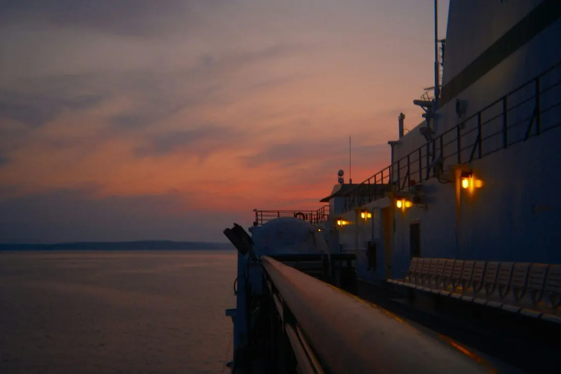 A ship's deck rail and glowing lights at dusk, with calm water under a pink and purple sky.