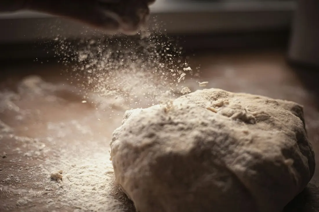Flour dusts a mound of bread dough as a hand sprinkles it from above.
