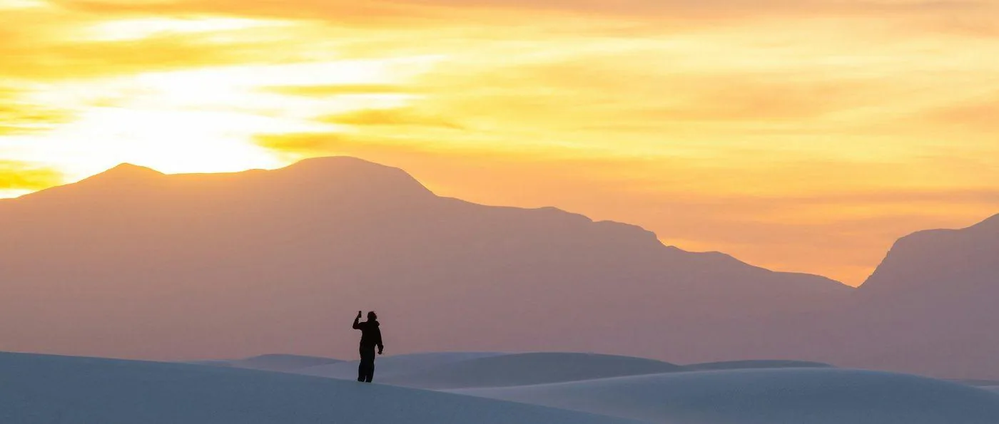 A traveler stands in silhouette on pale sand dunes at sunset, with layered mountains and a golden sky behind.