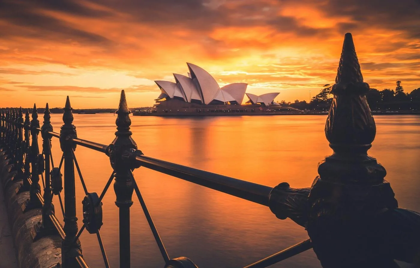 The Sydney Opera House glows across the harbor at sunset, framed by an iron railing.