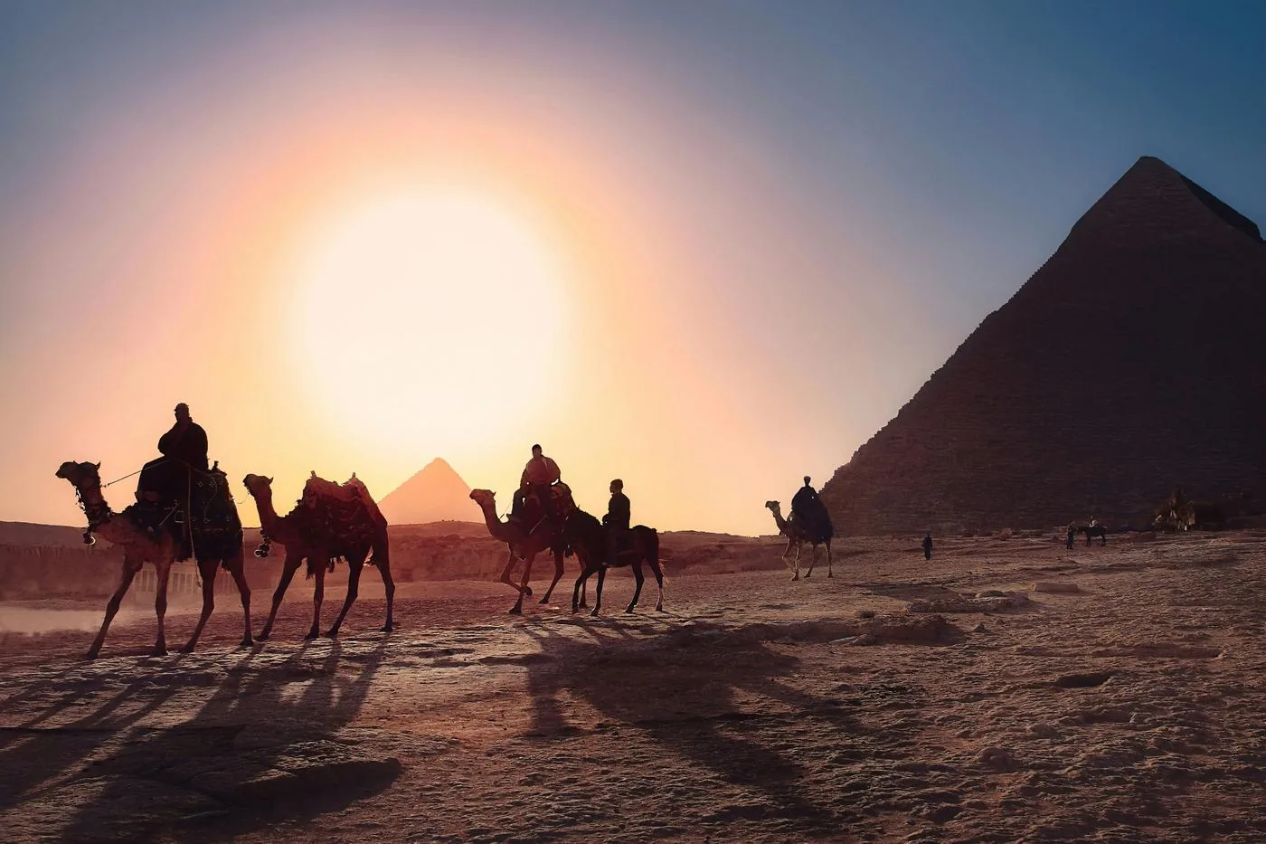 Camels and riders cross the desert in silhouette before a pyramid and a large setting sun.