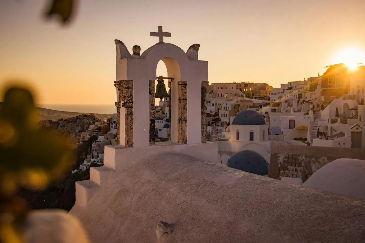 A white bell tower on Santorini frames whitewashed buildings and a blue-domed church at sunset.