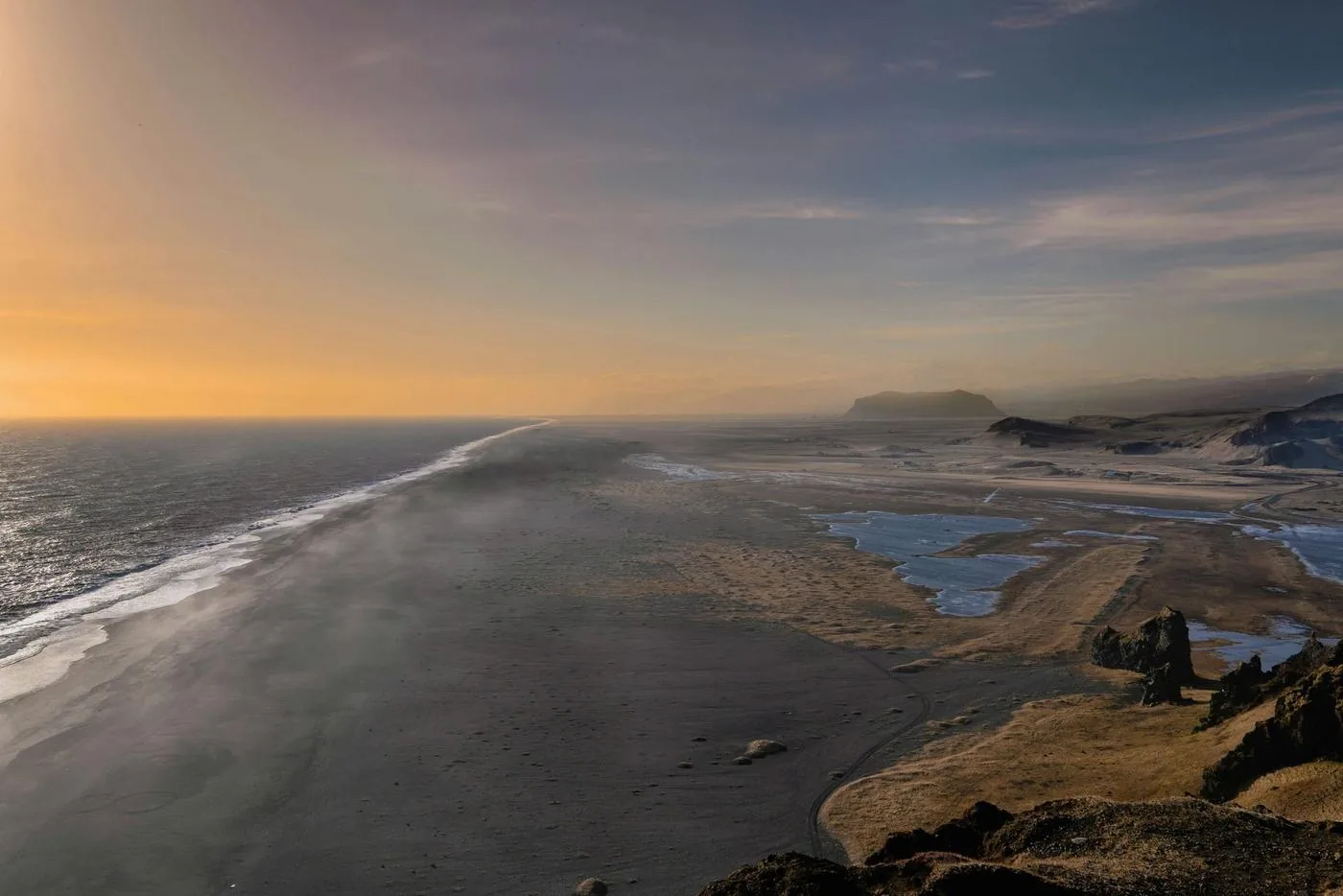 A black sand beach stretches along a windswept coast under a pale sunrise sky.