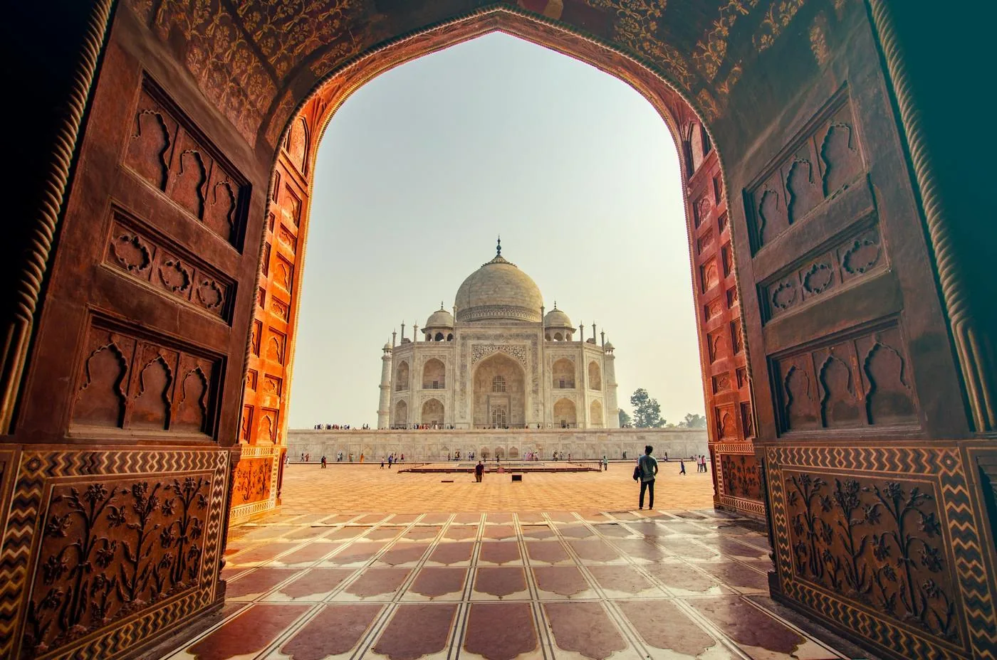 The Taj Mahal seen through a carved sandstone archway, with a lone visitor in the courtyard.