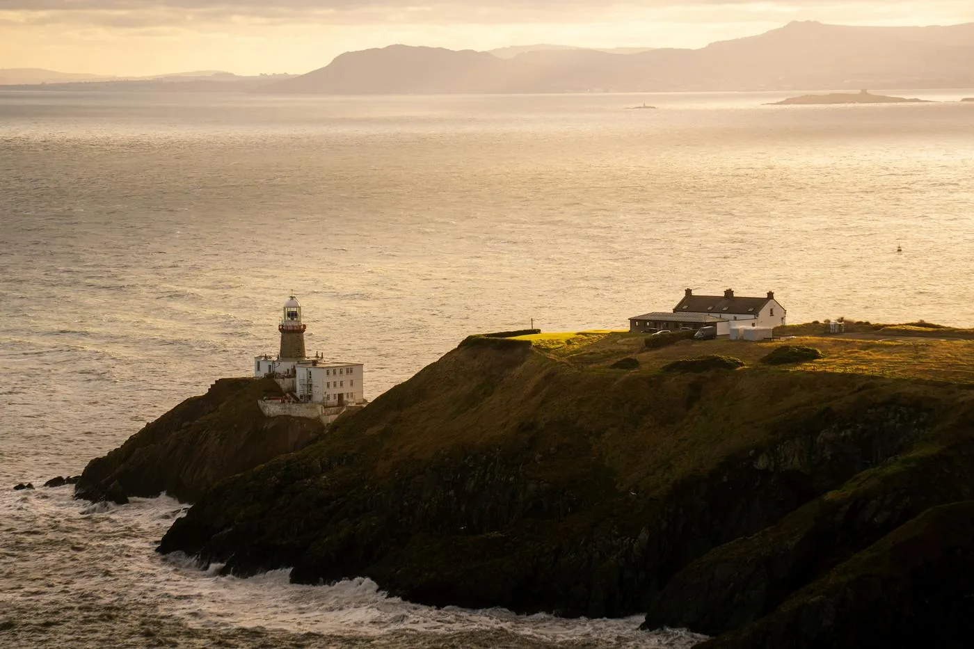 A lighthouse and a small white house sit on a green headland above calm sea in soft evening light.