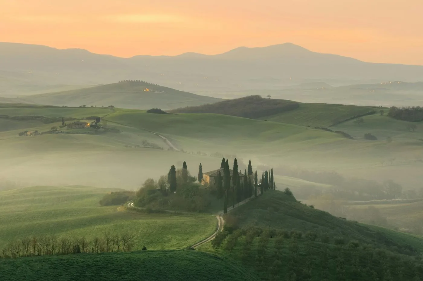 A farmhouse ringed by cypress trees on a green hill in morning mist, with rolling Tuscan countryside beyond.