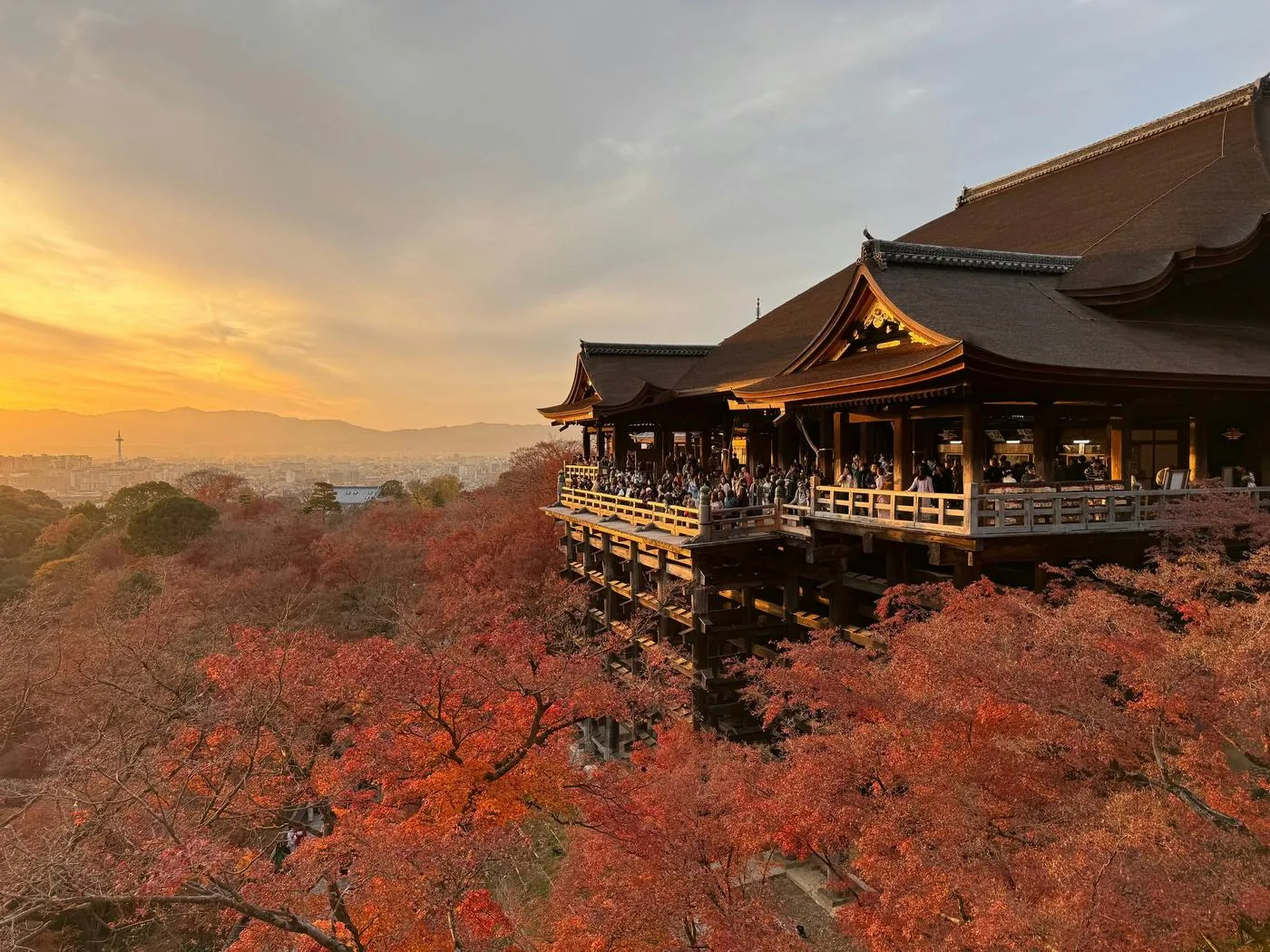 A wooden temple stage juts from a hillside above red and orange autumn maples at sunset.