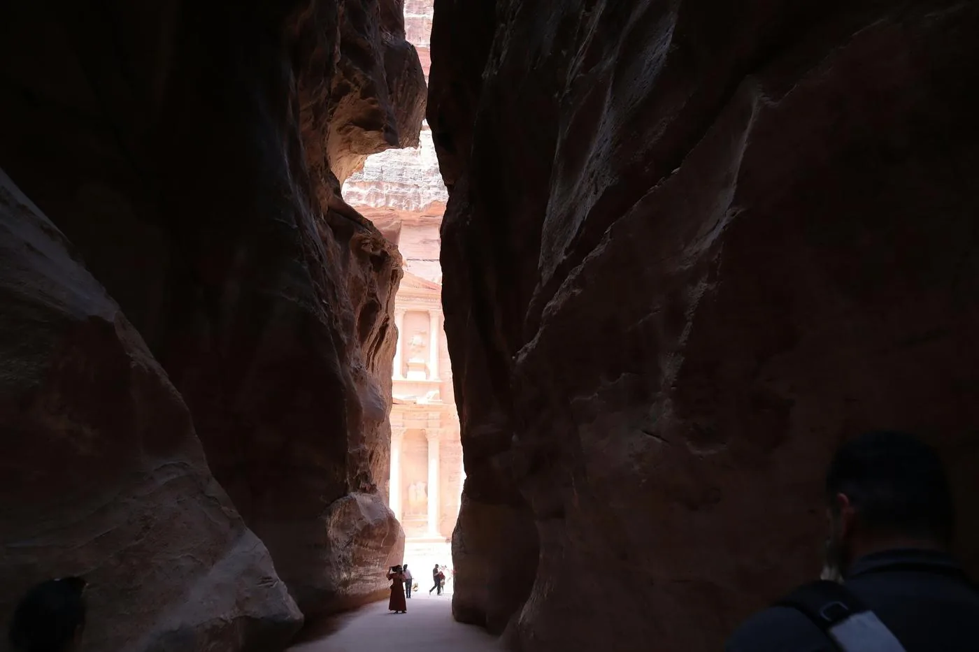 A narrow sandstone canyon opens onto the carved Treasury at Petra, with two small figures walking ahead.