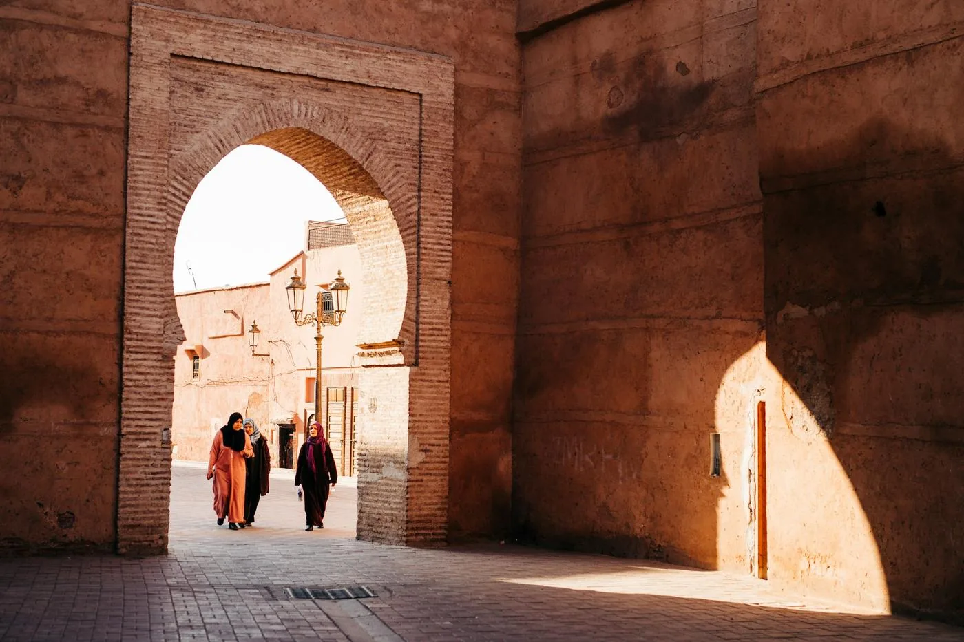 Two people walk through a tall brick archway in a warm, sunlit old city street.