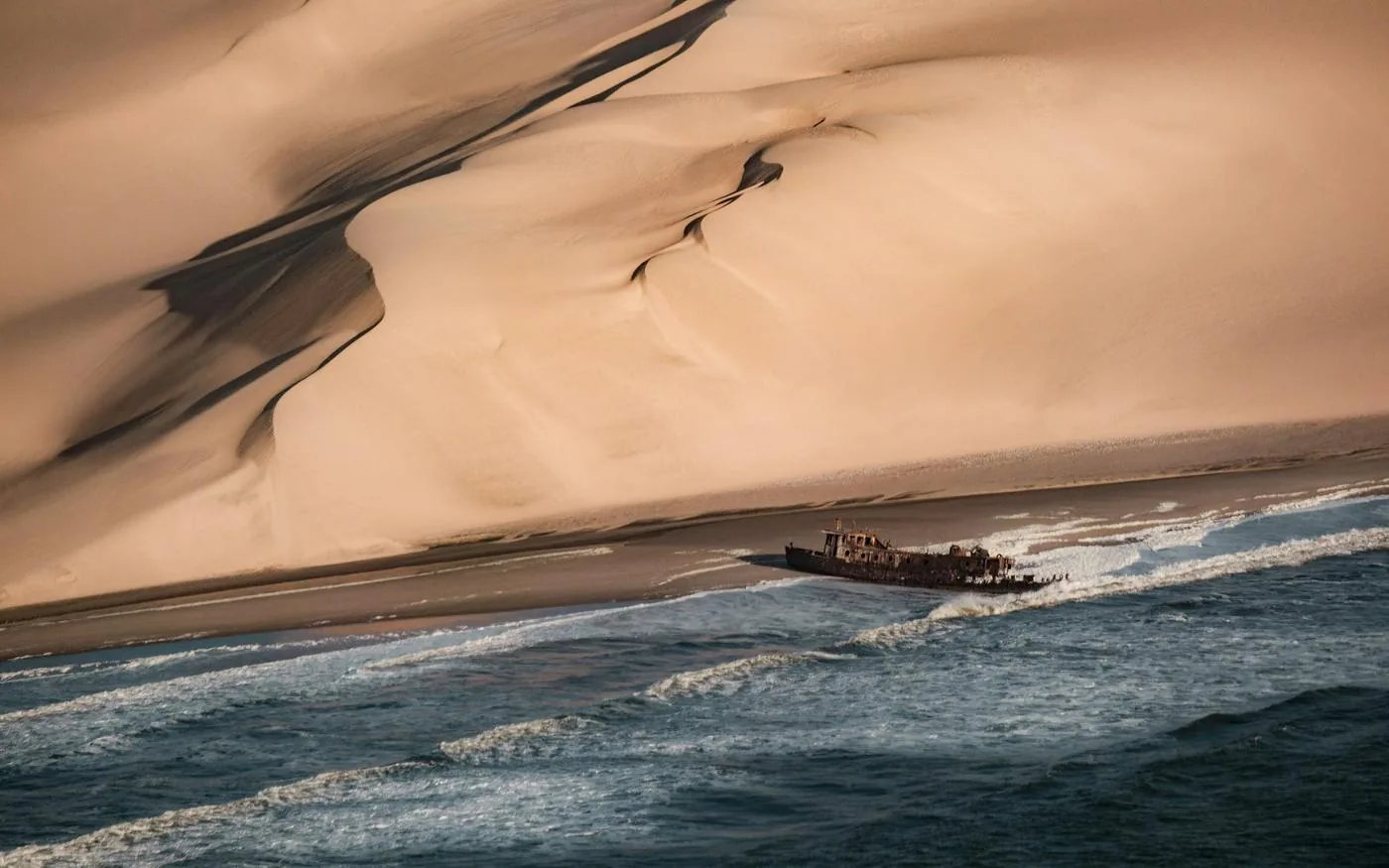 A rusted shipwreck in the surf below towering dunes on the Skeleton Coast, Namibia