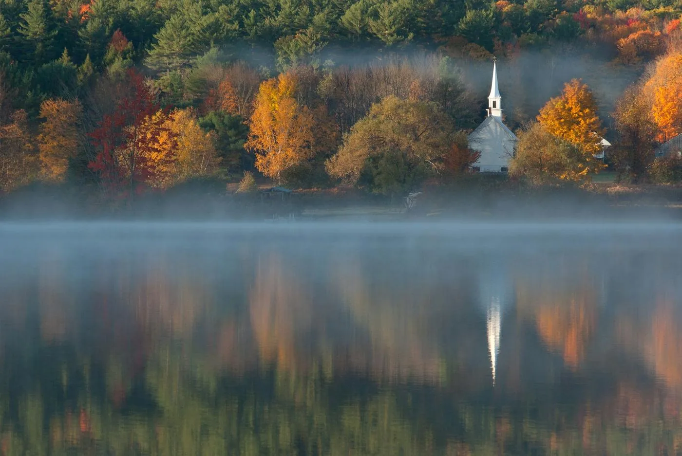 A white church steeple rises above autumn trees on a misty lakeshore, mirrored in the water.