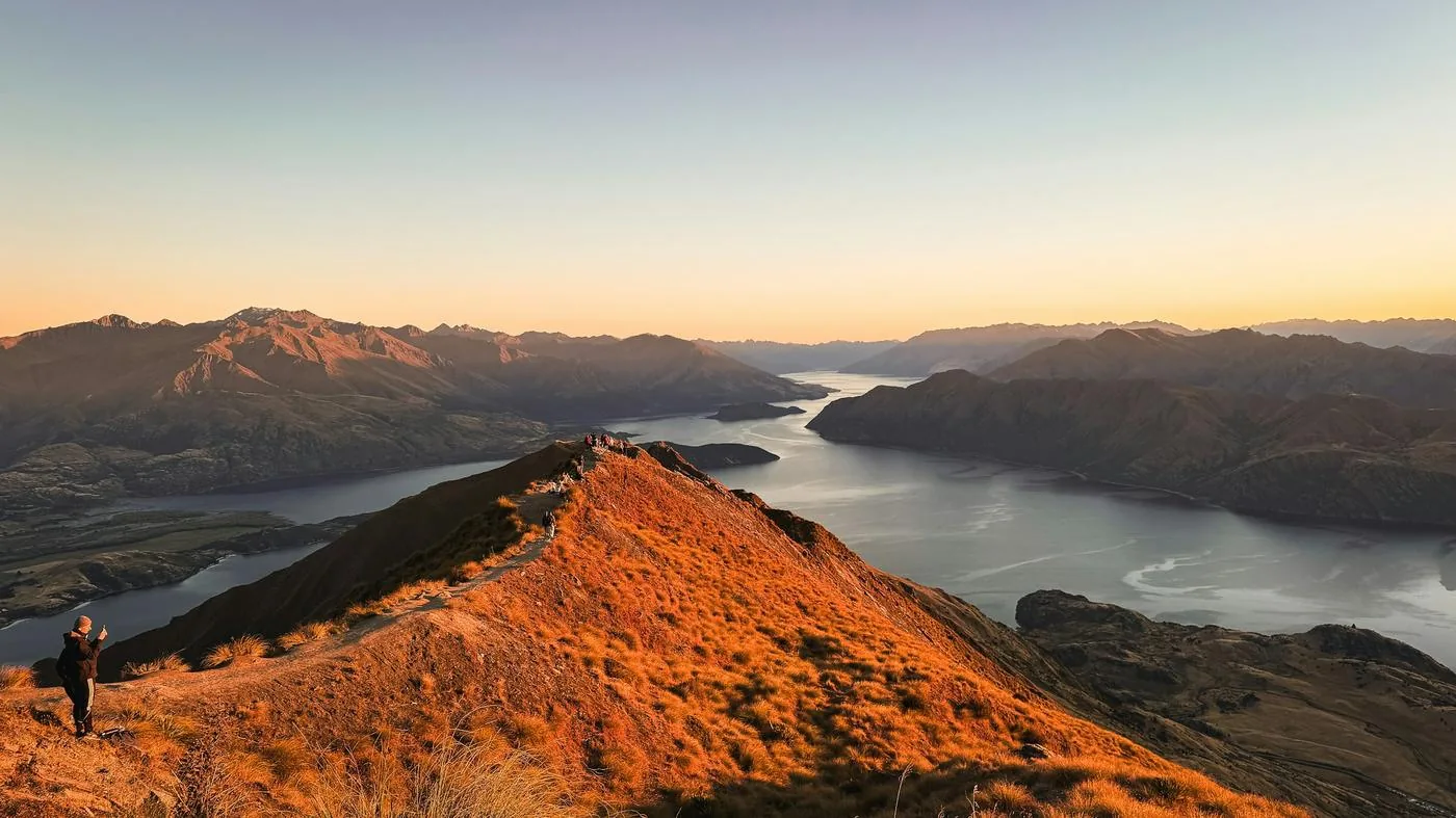 A hiker stands on an orange-lit ridge above a long blue lake and mountains at sunrise.
