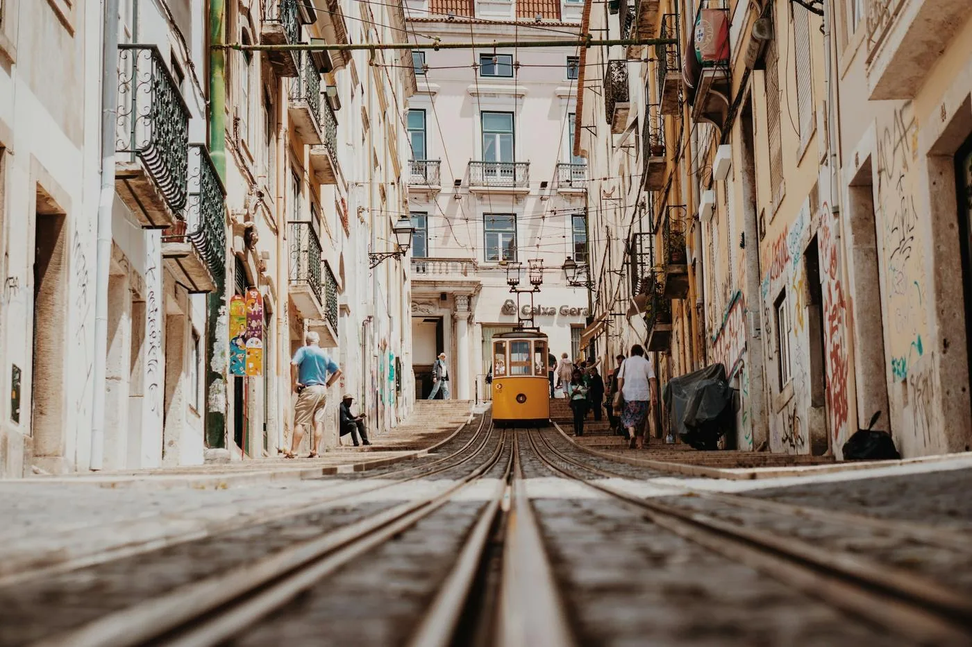 A yellow tram rolls up a narrow street between tall old buildings, seen low from the tracks.