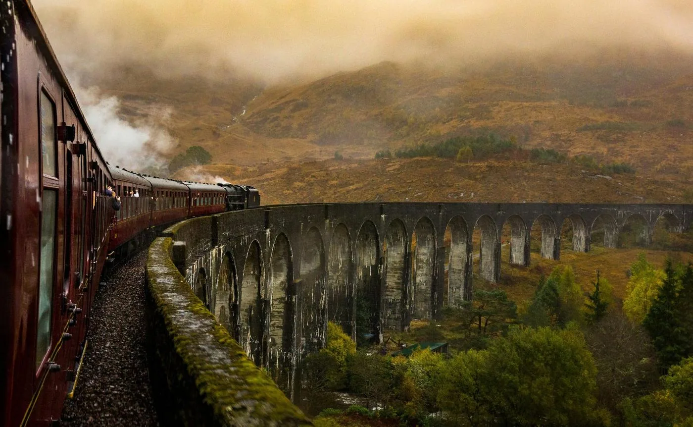 A vintage train crosses a curved stone viaduct through misty hills.