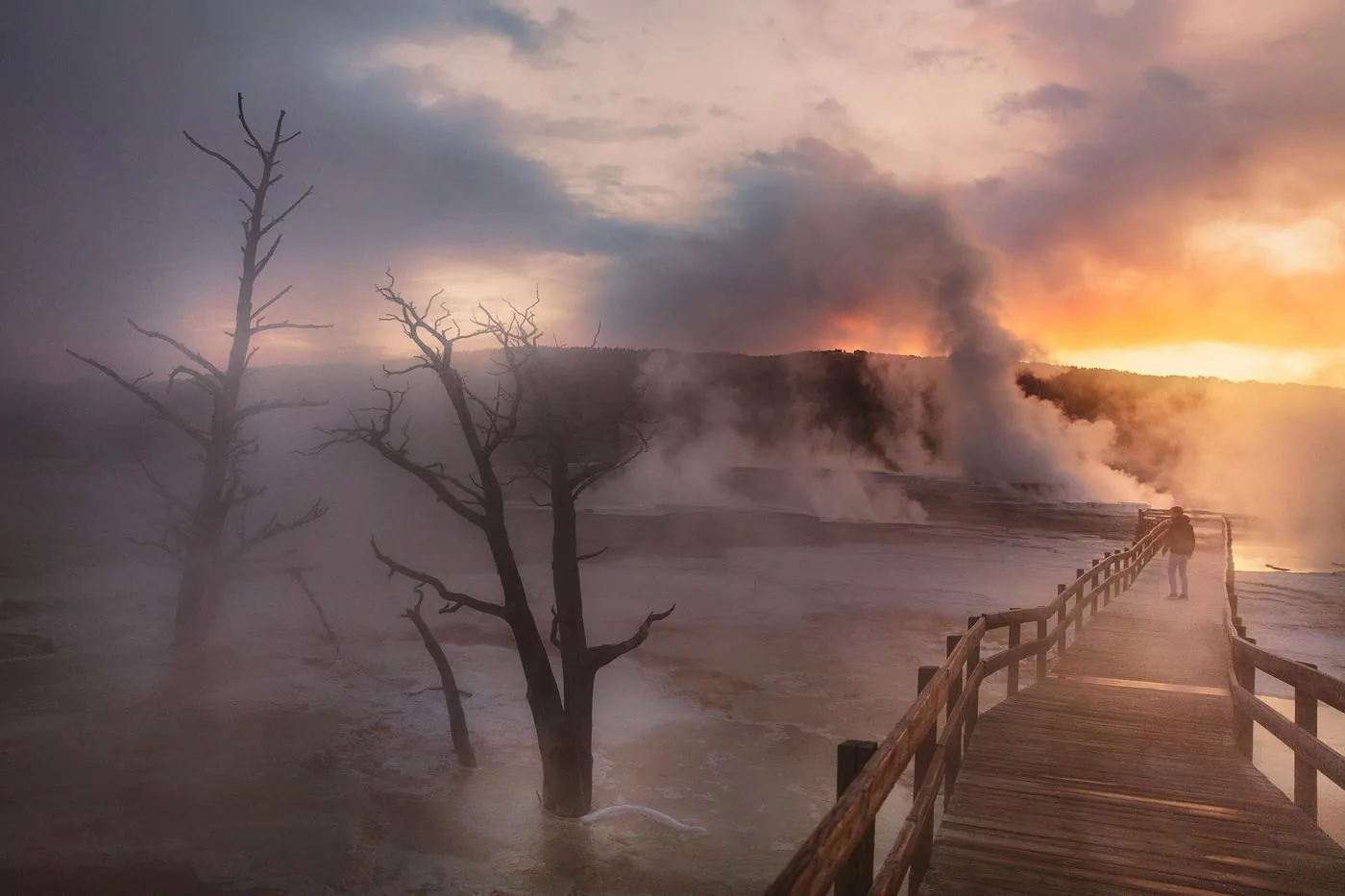 A boardwalk leads through steam and bare trees beside a geothermal pool under a stormy sunset sky.