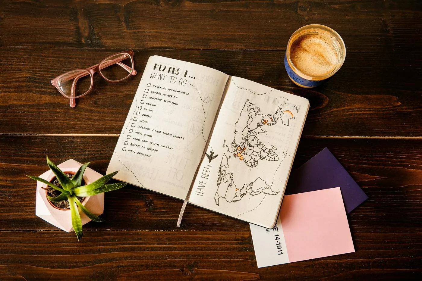 A travel journal open to a wish list of destinations and a hand-drawn world map, beside reading glasses, a coffee, and a small plant on a dark wooden table.